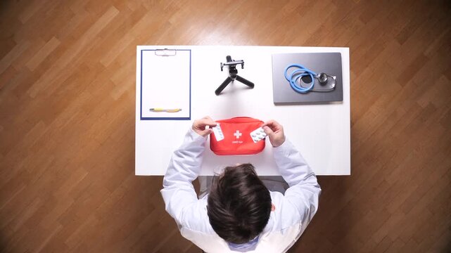 Doctor in lab coat at desk holding blister packs and opening a red first aid kit filled with medical supplies, top down view conveying preparation, care, and clinical professionalism