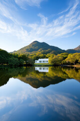 A serene landscape featuring a white building reflected in calm waters, surrounded by lush greenery and mountains under a blue sky with wispy clouds.