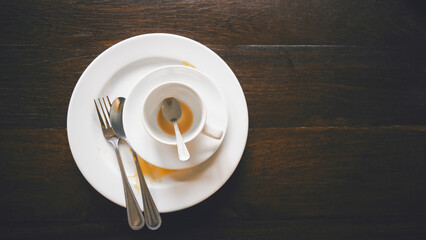 used white coffee cup and plate with spoon and fork on dark brown wooden desk after meal, top view