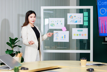 Asian confident businesswoman standing present marketing data work creative conference meeting room featuring chart display on glass board professional communication coworking modern office workplace.