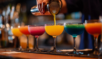 Bartender pouring vibrant layered cocktails into elegant glasses on bar counter, showcasing colorful drink presentation and smooth liquid flow