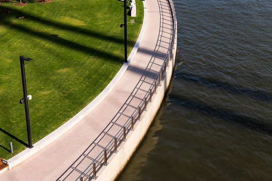 Looking down on curved river bank with path, fence, sharp shadows and green grass lawn