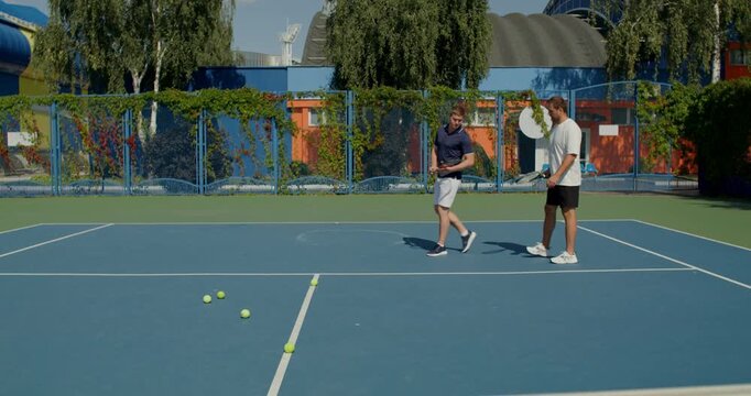 Two boys with rackets in their hands enter the tennis court . Two male tennis players train on the court with rackets in hand.