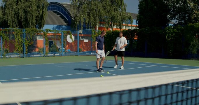 Two tennis players enter the court and begin practicing their swing. Two male tennis players train on the court with rackets in hand.
