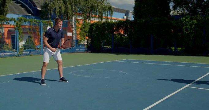 A guy talks while standing on the court, then starts hitting balls with his racket. Two male tennis players train on the court with rackets in hand.