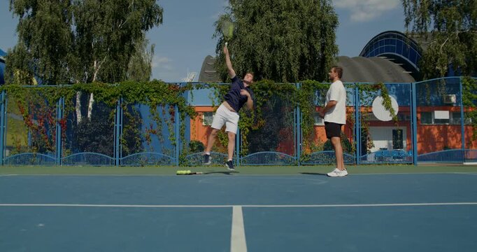 One guy serves balls to another, who practices hitting them with a racket on the court. Two male tennis players train on the court with rackets in hand.