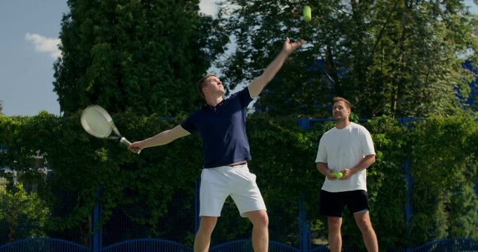 A guy hits the ball, then serves with a racket. Two male tennis players train on the court with rackets in hand.