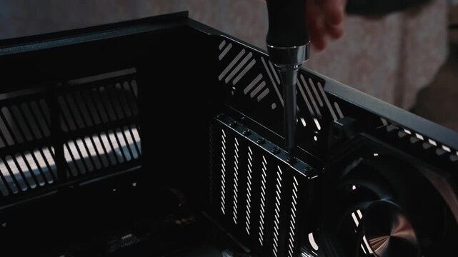 Unscrewing back panel screws of computer case before build, close-up of hand with screwdriver removing fasteners near the fan inside black chassis
