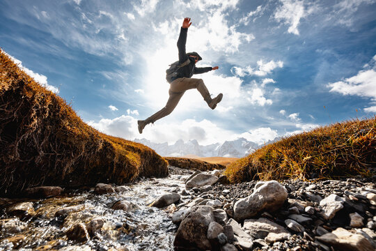 Man hiker with backpack jumps over small creek against high snow capped mountains. Active vacations concept - Powered by Adobe