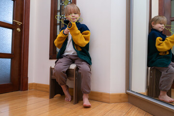 Little boy sitting on a stool holding a green hanger. Kid playing with clothes near mirror © Victor zastol'skiy