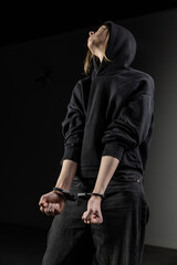 A young woman in a black hoodie stands handcuffed with her hands in front, looking to the side. The dim lighting and her posture convey tension and introspection © ugis