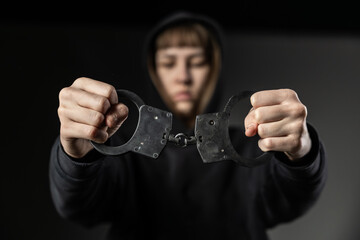 A young person in a hoodie holds a pair of metal handcuffs forward with a serious expression, creating a powerful, confrontational mood. Dark background adds emphasis