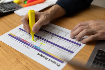 Person highlighting figures on project budget sheet with yellow marker, analyzing financial data and expenses. Concept for budgeting, accounting, financial planning business cost control.
