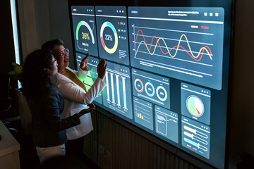 Young Indian and senior Asian woman review interactive charts on a large digital screen in a modern office, showcasing corporate diversity, tech leadership, data analysis, and collaborative strategy.