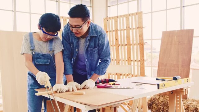 Asian father teaches woodworking to his son in a bright workshop. Both wear safety gear and work with tools at a wooden table, sharing skills, patience, and hands-on learning time together.
