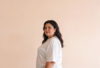 An overweight adult woman in her 30s with long dark hair stands in profile wearing a white t-shirt against a plain light peach background, looking directly at the camera