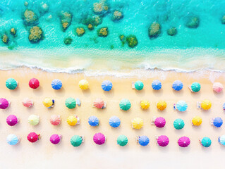 Brightly colored umbrellas spread across a sandy beach, providing shade to visitors enjoying the warm sun. Crystal-clear waters gently wash ashore, creating a tropical paradise atmosphere.