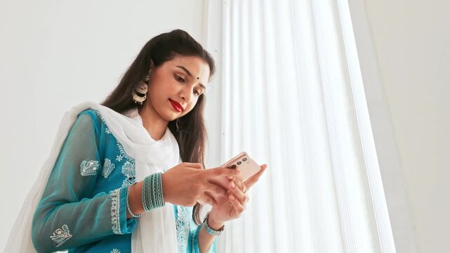 A woman in traditional Indian attire talks on the phone near a bright window. She wears a blue and white salwar kameez with elegant jewelry, engaged in a conversation at home. Real time 4k video.