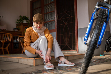 Asian muslim woman sits on a doorstep adjusting her sandals, wearing a hijab and cap, with a blue bicycle beside her in a cozy home setting.