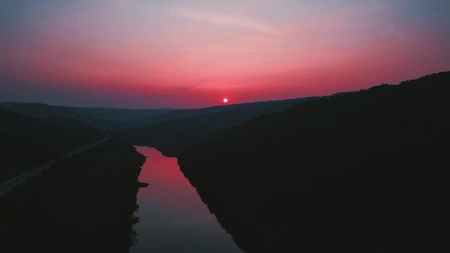 Cinematic slow backward and down shot of a small river at dusk, reddish sunset reflections on calm water, dark serene mood with a parallel highway adding subtle urban contrast.