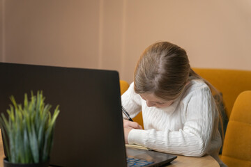 Young girl concentrating on homework at a table with laptop and notebook, writing notes during...