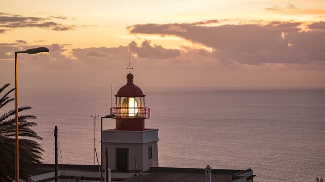 Timelapse close-up of the Farol da Ponta do Pargo lighthouse after sunset on Madeira Island, Portugal. The lighthouse beacon rotates and casts powerful light beams across the sky while soft sunset clo