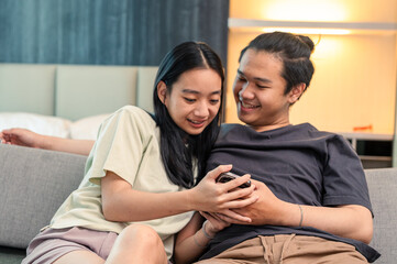 Asian couple sitting close together on a couch smiling while looking at a smartphone, enjoying a funny moment and bonding happily in a cozy living room with warm lighting