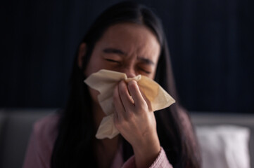 Asian woman in pink pajamas sneezing into a tissue while sitting on bed, looking unwell with signs of cold or allergy in a dim bedroom
