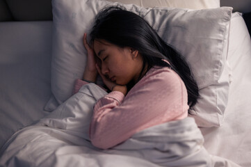 An Asian woman in pink pajamas sleeping on her side in bed with white pillows and blanket, resting her head on her hand, showing peaceful relaxation and comfort in a cozy bedroom scene