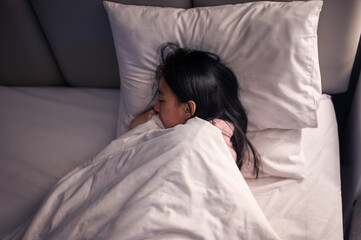 An Asian woman in pink pajamas sleeping on her side in bed with white pillows and blanket, resting her head on her hand, showing peaceful relaxation and comfort in a cozy bedroom scene