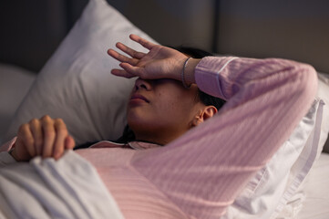 An Asian woman in pink pajamas lying in bed with white pillows and blanket, covering part of her face with her arm, showing tiredness, stress, or discomfort in a bedroom setting