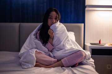 Asian woman sitting on bed wrapped in a white blanket, wearing pink pajamas and looking tired and unwell in a dimly lit bedroom environment