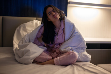 Asian woman sitting on bed wearing pink pajamas and covered with a white blanket, holding her stomach and looking unwell in a dimly lit bedroom