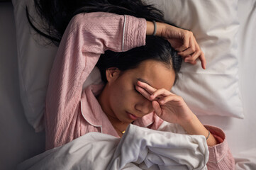 An Asian woman in pink pajamas lying in bed with white pillows and blanket, covering part of her face with her arm, showing tiredness, stress, or discomfort in a bedroom setting