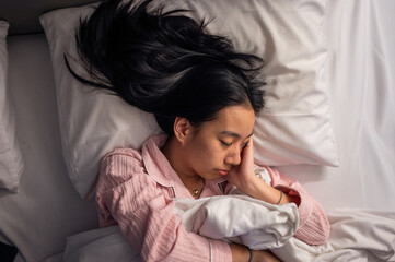 An Asian woman in pink pajamas sleeping on her side in bed with white pillows and blanket, resting her head on her hand, showing peaceful relaxation and comfort in a cozy bedroom scene