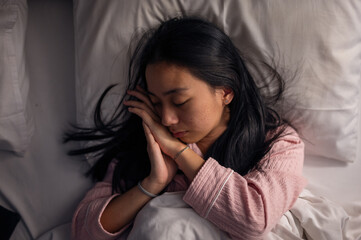 An Asian woman in pink pajamas sleeping peacefully on a bed with white pillows and blanket, eyes closed, resting on her side, showing calmness, relaxation, and comfort in a bedroom scene