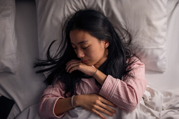 An Asian woman in pink pajamas sleeping peacefully on a bed with white pillows and blanket, eyes closed, resting on her side, showing calmness, relaxation, and comfort in a bedroom scene