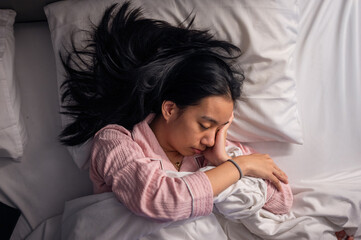 An Asian woman in pink pajamas sleeping on her side in bed with white pillows and blanket, resting her head on her hand, showing peaceful relaxation and comfort in a cozy bedroom scene