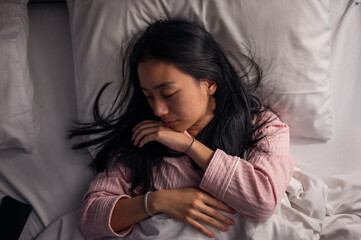 An Asian woman in pink pajamas sleeping peacefully on a bed with white pillows and blanket, eyes closed, resting on her side, showing calmness, relaxation, and comfort in a bedroom scene