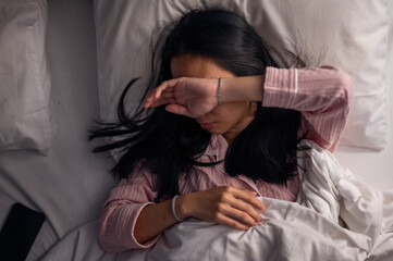 An Asian woman in pink pajamas lying in bed with white pillows and blanket, covering part of her face with her arm, showing tiredness, stress, or discomfort in a bedroom setting