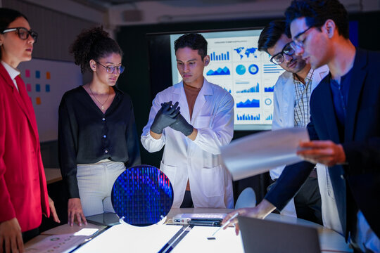 Specialist team of engineers and executives inspecting a large silicon wafer in a high-tech semiconductor laboratory. Discussion on microchip and RAM memory manufacturing process and quality control. - Powered by Adobe