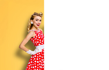 Beautiful expressive smiling woman wear pinup red dress pointing white blank banner with mockup free text space. Pin up girl advertise, isolated yellow background. Retro vintage sales ad image
