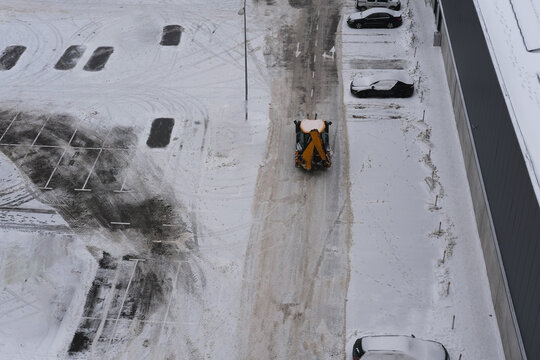 Snow plow clearing a parking lot covered in fresh snow, with tire tracks visible and parked vehicles in the background, showcasing winter maintenance and urban landscape