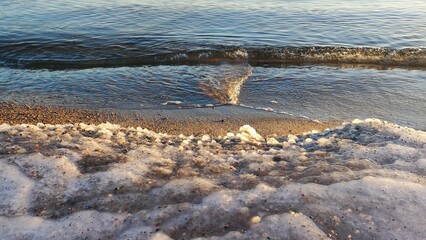 Snowy Shoreline and Freezing Sea. A gentle wave rolls onto a snowy beach, creating a serene contrast between water and snow. The sunset casts a warm light over the scene.
