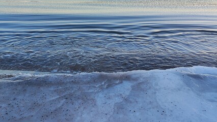 Snowy Shoreline and Freezing Sea. A gentle wave rolls onto a snowy beach, creating a serene contrast between water and snow. The sunset casts a warm light over the scene.