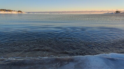 Calm Coastal Morning and Freezing Sea. Gentle waves roll onto the shoreline under a serene sky. Distant landforms provide a picturesque backdrop as the sun rises.