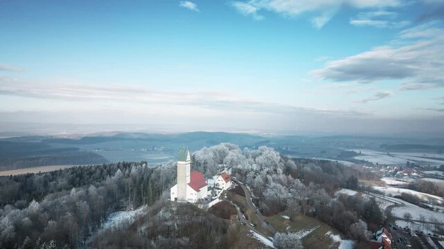 Drone performs a smooth clockwise orbit around the prominent white tower chapel on the hilltop in Utterweiler, Baden-W&uuml;rttemberg, showcasing expansive frosty forests, snowy meadows, and distant valley