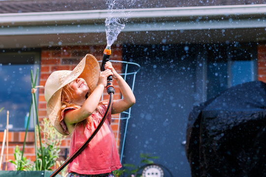 Little girl playing outdoors with hose in backyard wearing big floppy sun hat - water play