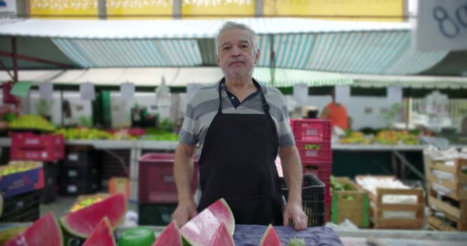 Elderly market vendor standing behind produce stall with sliced watermelon, everyday work routine, local food market life, authenticity, community presence