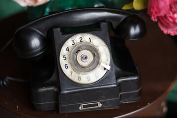 Vintage black rotary telephone with a classic design, featuring a circular dial and a coiled cord, placed on a wooden table surrounded by colorful flowers, evoking nostalgia and charm